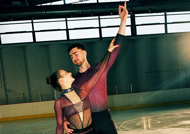 Eiskunstläufer Robert Kunkel und Annika Hocke stehen Rücken an Rücken in schwarz, weiß, grauer Athleisure und lächeln sanft (Foto)